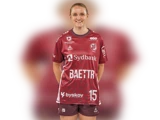 En kvinde i en rødbrun sportsdragt med nummer 15 står smilende med hænderne bag ryggen foran en sløret hvid baggrund.